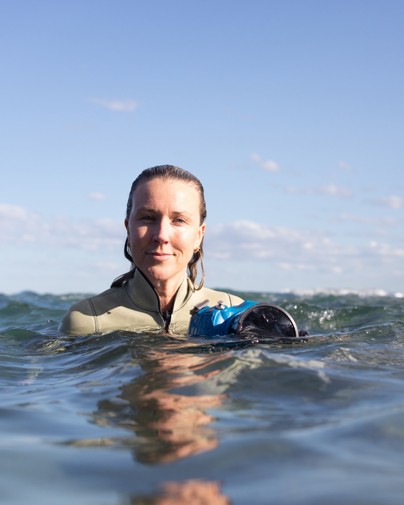 Woman in wetsuit holding a camera underwater with clear blue sky and ocean water.