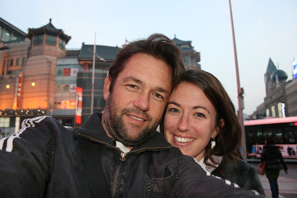 Two people taking a selfie in an urban setting with buildings and a bus in the background.