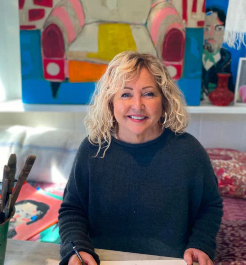 Woman with curly blonde hair wearing a dark sweater in a room with colorful artwork on the wall.
