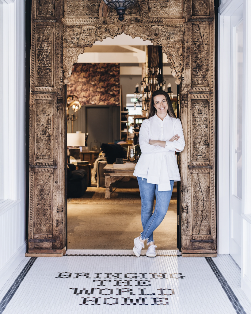 Woman standing in a doorway with ornate wooden frame, interior decor visible.
