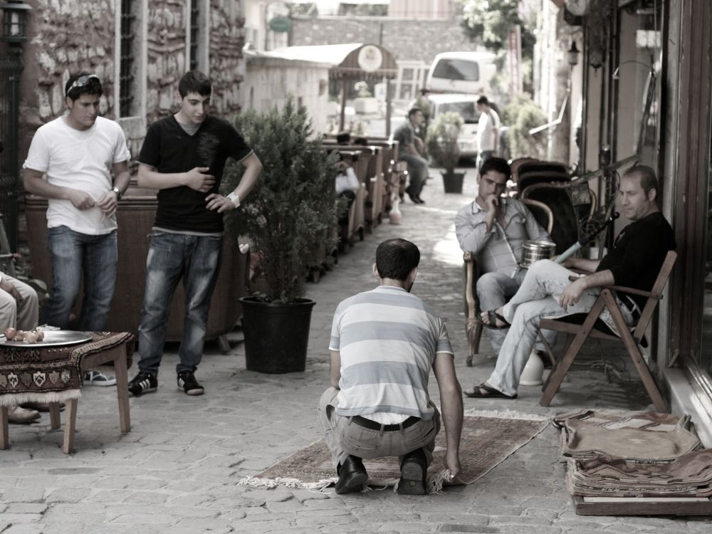 People gathered in an outdoor space in Turkey with a man kneeling on the ground displaying a vintage Turkish rug.